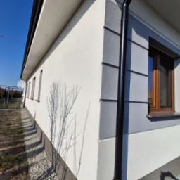 Świeżo pomalowana elewacja domu jednorodzinnego w odcieniach szarości i bieli, z widoczną rynną, oknem z drewnianą ramą i cieniem drzewa na ścianie.