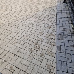 Dariusz Lewandowski - Częściowo umyta kostka brukowa ułożona w jodełkę, widoczne różnice w kolorze. Po prawej stronie kostka ciemniejsza, po lewej jaśniejsza. Fragment metalowego ogrodzenia.