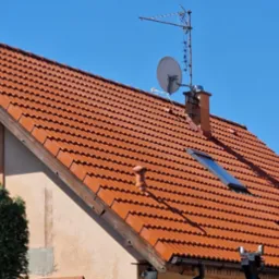 Czerwona dachówka karpiówka na skośnym dachu domu jednorodzinnego, widoczny komin, okno dachowe i antena satelitarna na tle błękitnego nieba.