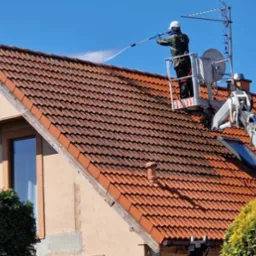 Pracownik na podnośniku czyści dach z czerwonej dachówki za pomocą myjki ciśnieniowej. Widoczna różnica między umytą, a brudną częścią dachu. Błękitne niebo w tle.