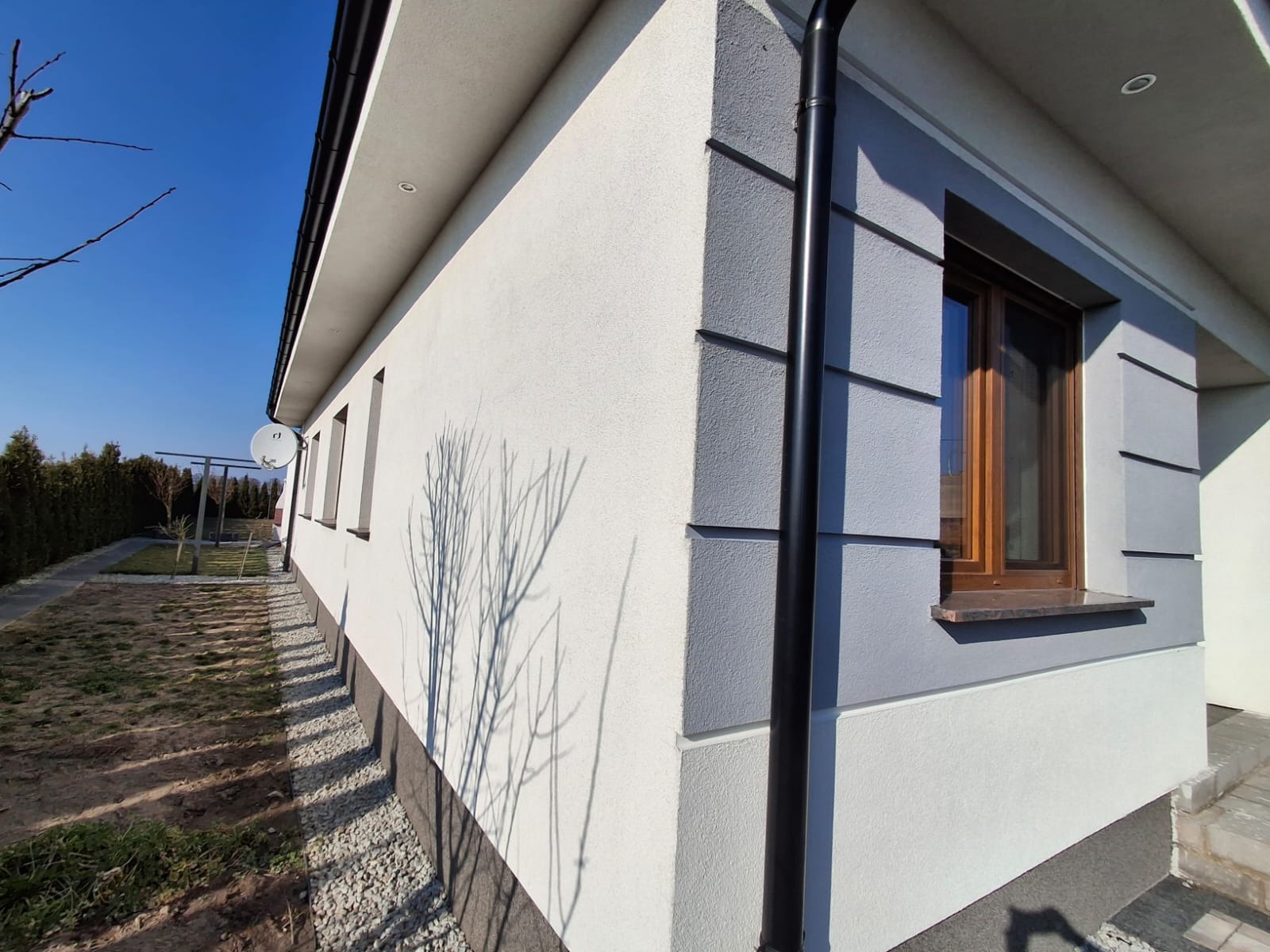 Świeżo pomalowana elewacja domu jednorodzinnego w odcieniach szarości i bieli, z widoczną rynną, oknem z drewnianą ramą i cieniem drzewa na ścianie.