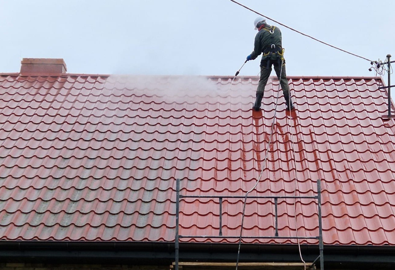 Czerwony dach z dachówki ceramicznej czyszczony myjką ciśnieniową przez pracownika w kombinezonie ochronnym i kasku. Widoczny komin i drabina dekarska.