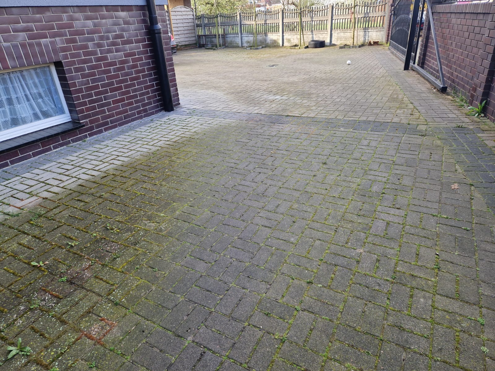 Częściowo wyczyszczona kostka brukowa przed domem, z widoczną różnicą w kolorze i brakiem mchu po lewej stronie. Dom z cegły i brama w tle.