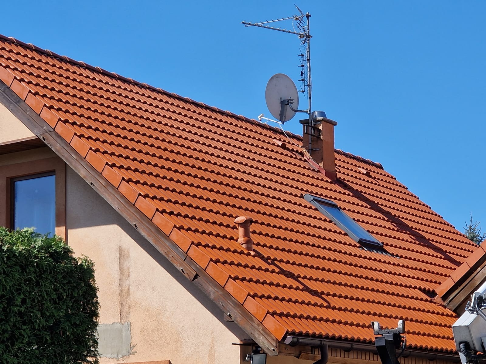 Czerwona dachówka karpiówka na skośnym dachu domu jednorodzinnego, widoczny komin, okno dachowe i antena satelitarna na tle błękitnego nieba.