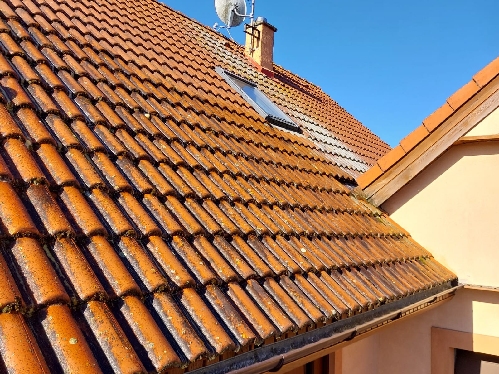 Ceglasty dach z dachówką ceramiczną, pokryty mchem, widoczny z bliska pod kątem, z lukarną, kominem i anteną satelitarną na tle błękitnego nieba.