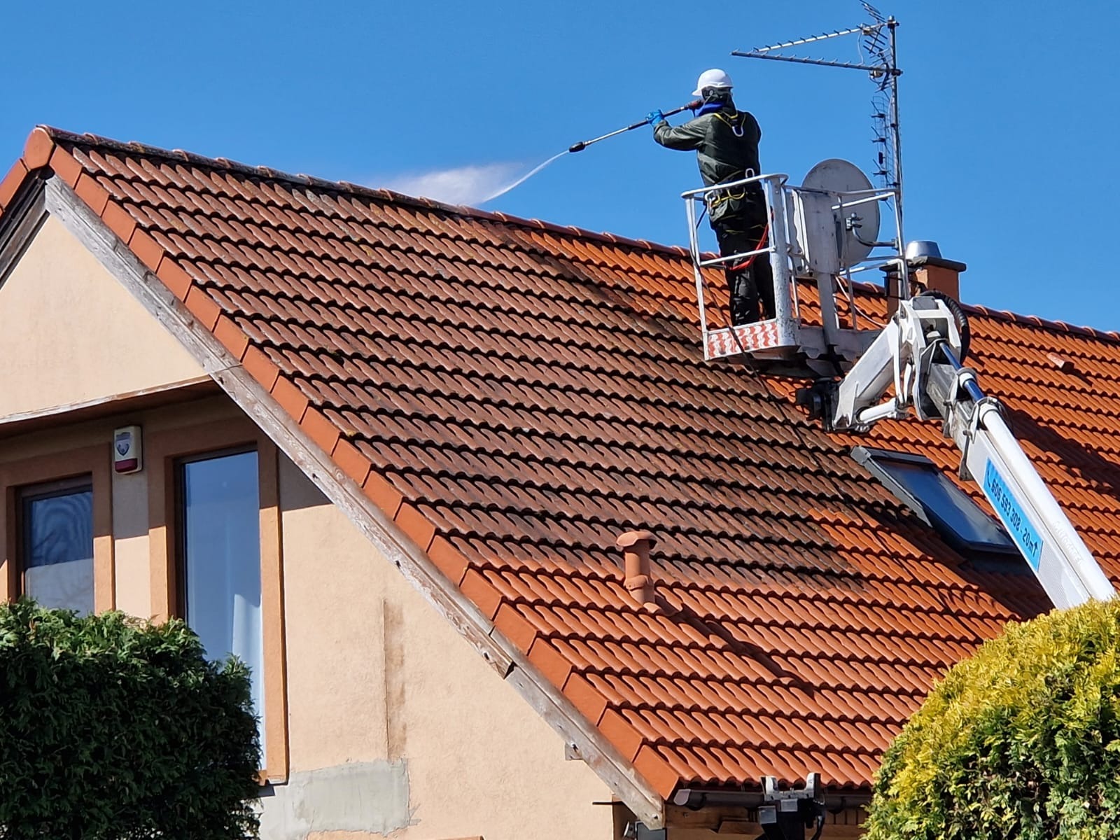 Pracownik na podnośniku czyści dach z czerwonej dachówki za pomocą myjki ciśnieniowej. Widoczna różnica między umytą, a brudną częścią dachu. Błękitne niebo w tle.