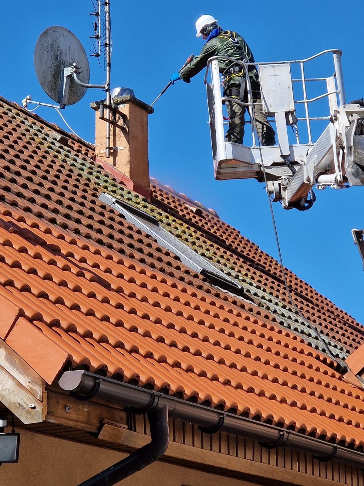 Pracownik w koszu podnośnika myje ciśnieniowo dach z czerwonej dachówki, z widocznym kominem i anteną satelitarną, fragment dachu umyty, niebieskie niebo w tle.
