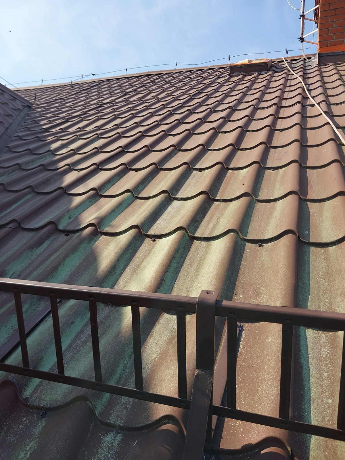 Miedziany dach z metalowej dachówki, częściowo pokryty zielonym nalotem, widziany z dołu pod kątem. Fragment barierki w dolnej części kadru. Słoneczny dzień.