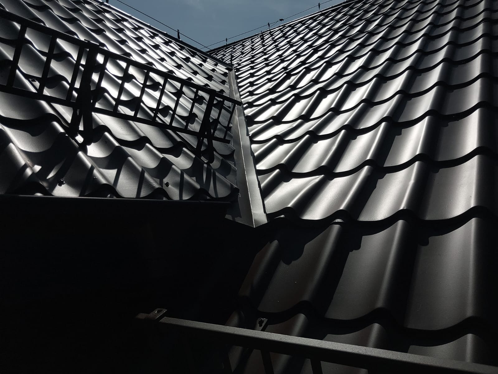 Błyszczący, czarny dach z metalowej dachówki, widoczny z dołu pod kątem. Fragment barierki i rynny. Niebo w tle.