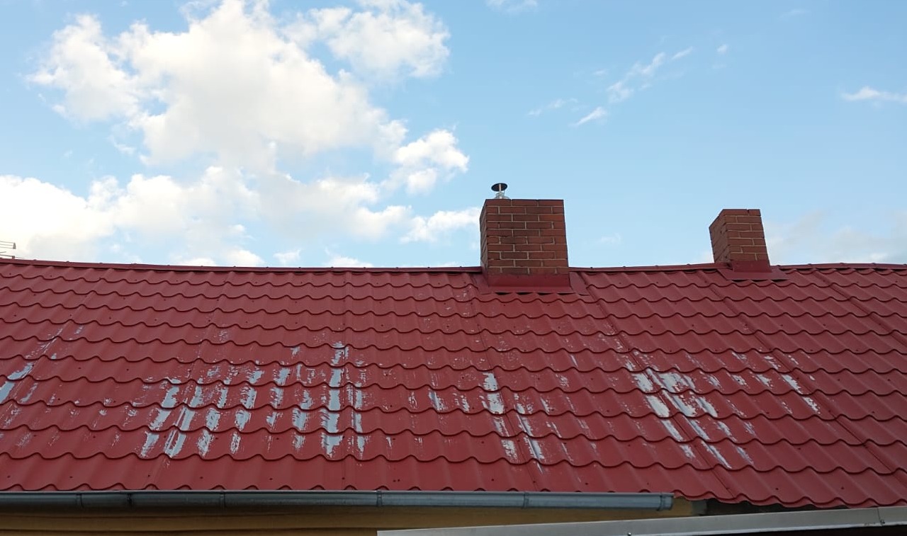 Czerwony dach z blachodachówki z dwoma kominami z cegły na tle błękitnego nieba z chmurami. Widoczne ślady wcześniejszych prac na dachu.