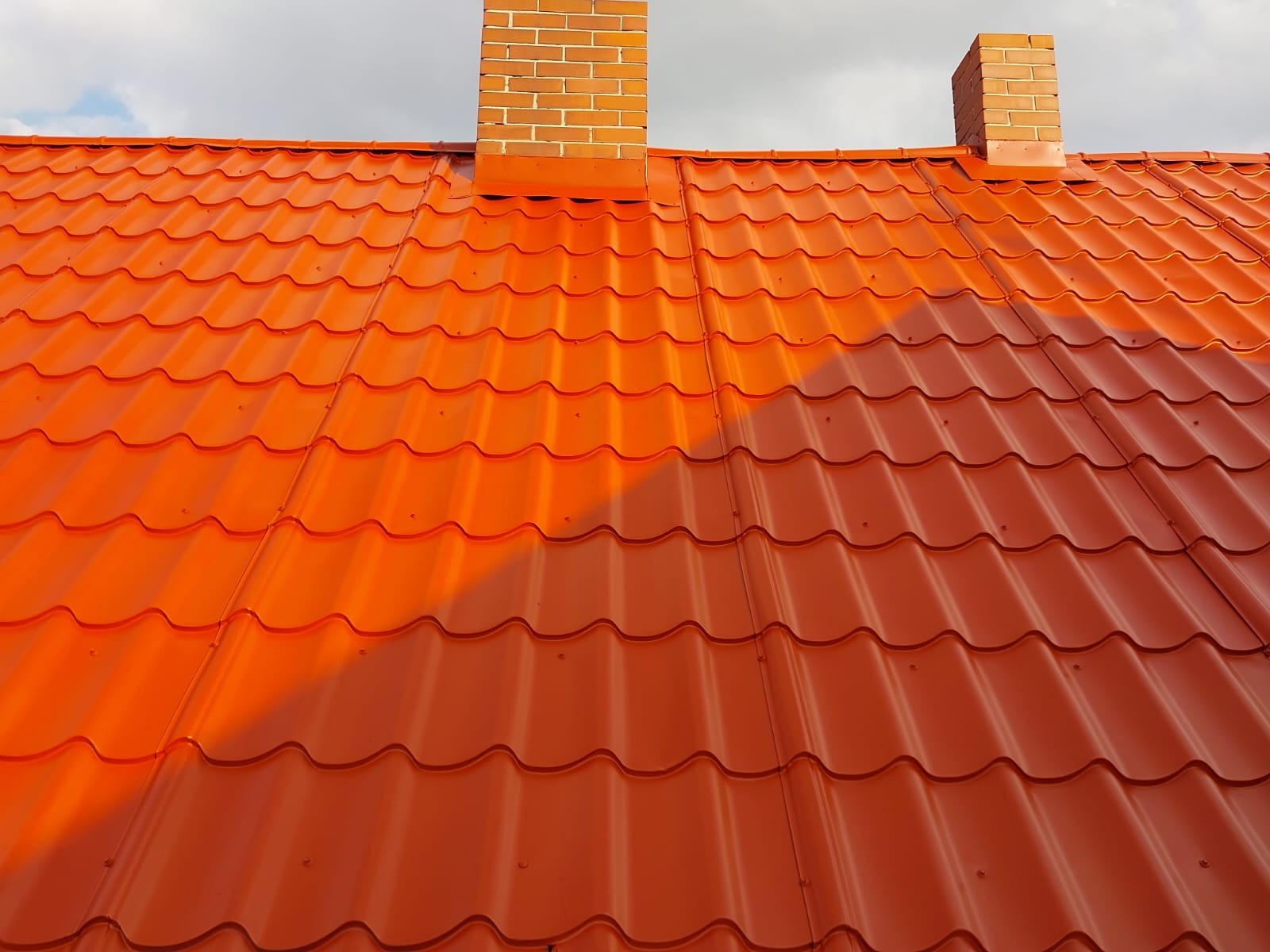 Czerwona blachodachówka z dwoma kominami z cegły w ciepłym świetle słonecznym. Widok z dołu na dach z wyraźnym wzorem fal i widocznymi mocowaniami.
