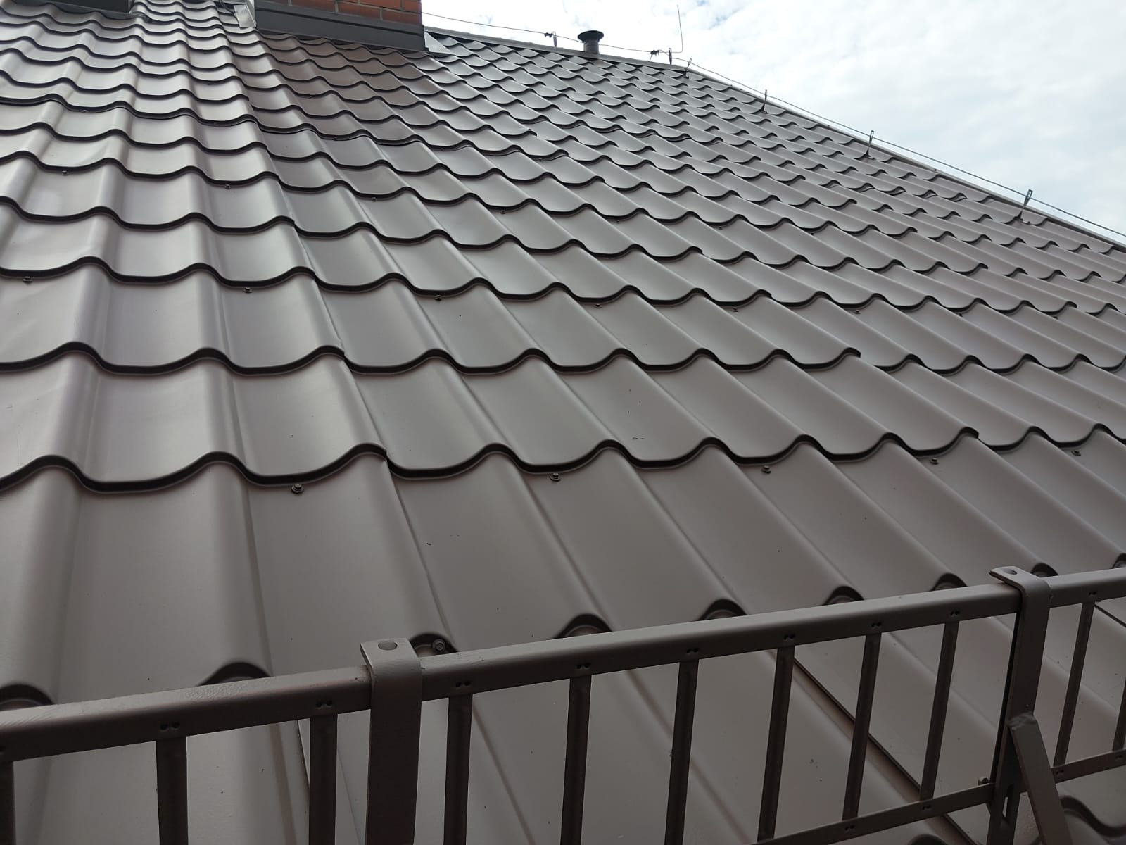 Ciemnobrązowy dach z blachodachówki modułowej, widoczny z dołu pod kątem. W tle fragment komina i niebo. Na pierwszym planie fragment metalowej barierki.