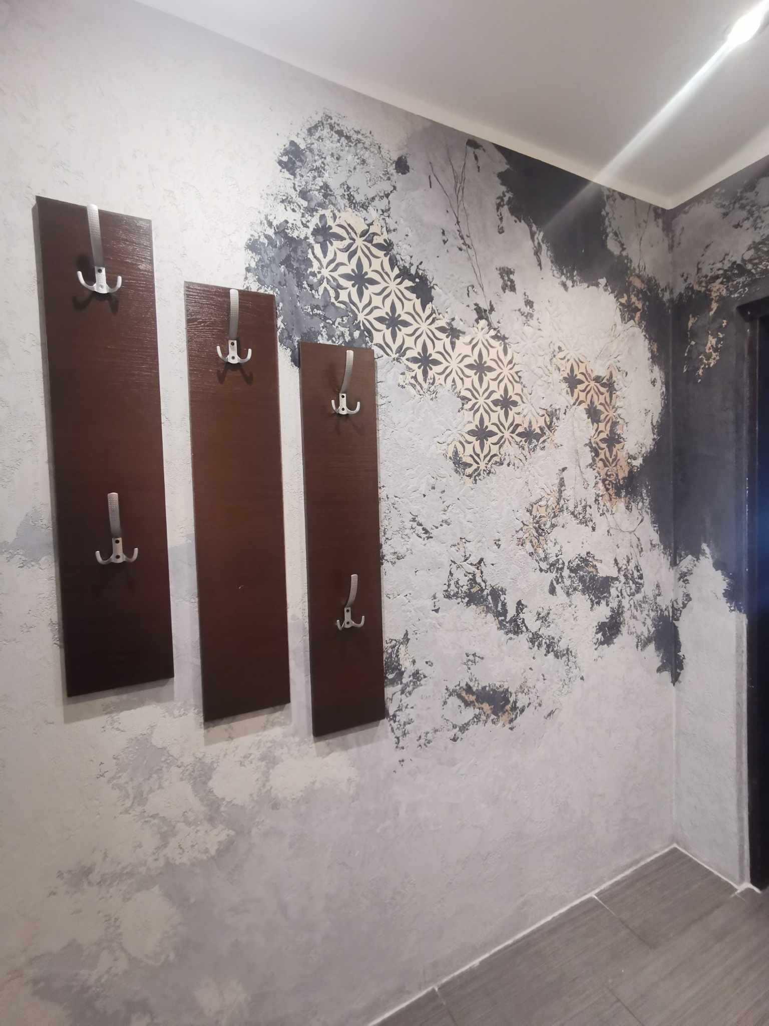 Ściana z dekoracyjnym tynkiem imitującym marmur w odcieniach szarości i beżu, uzupełniona o wzór geometryczny i trzy drewniane panele z metalowymi wieszakami.