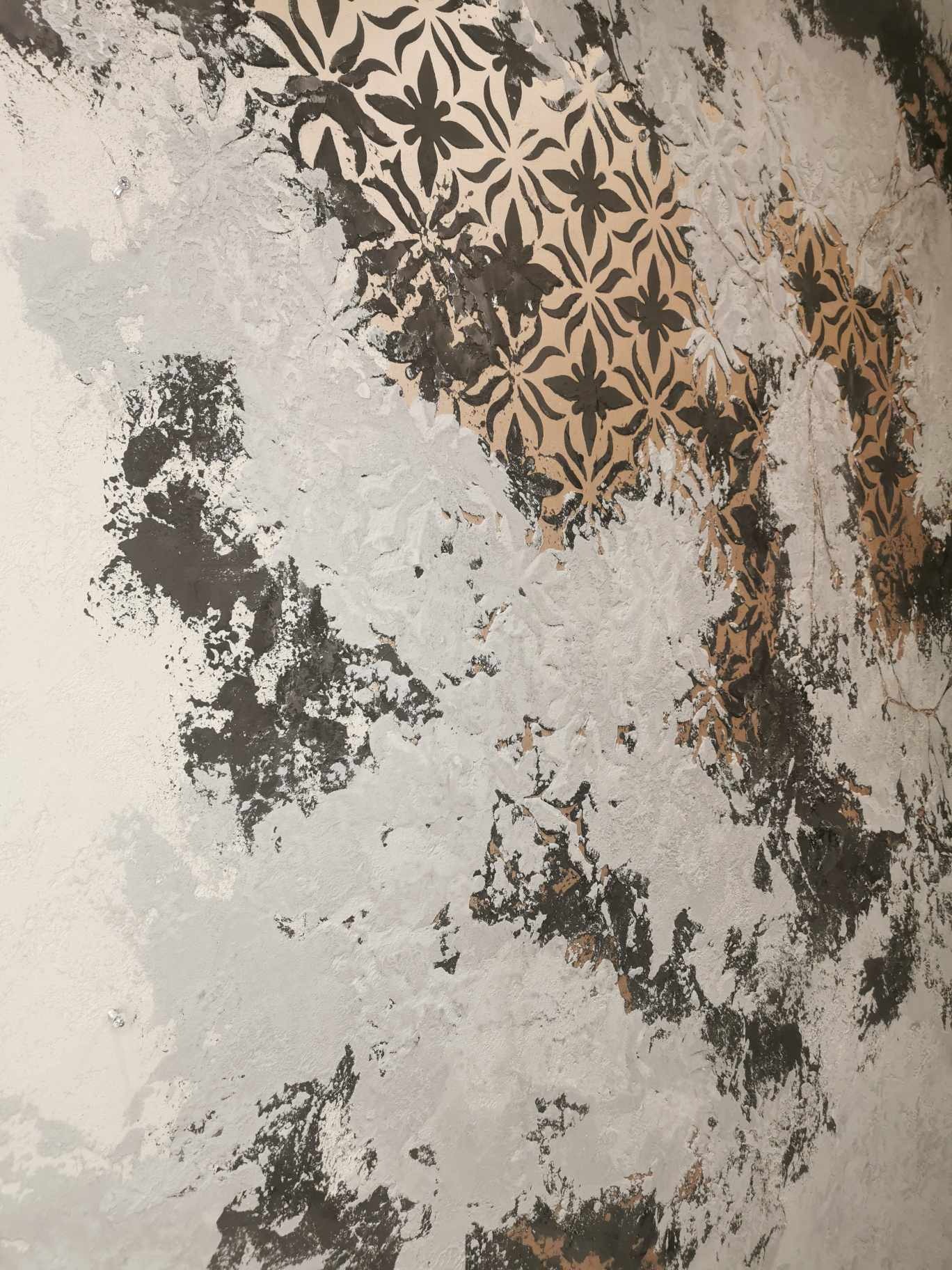 Nierówna powierzchnia ściany z fragmentami dekoracyjnego wzoru kwiatowego w kolorach beżu i ciemnej szarości, częściowo pokryta jasną farbą z efektem struktury.