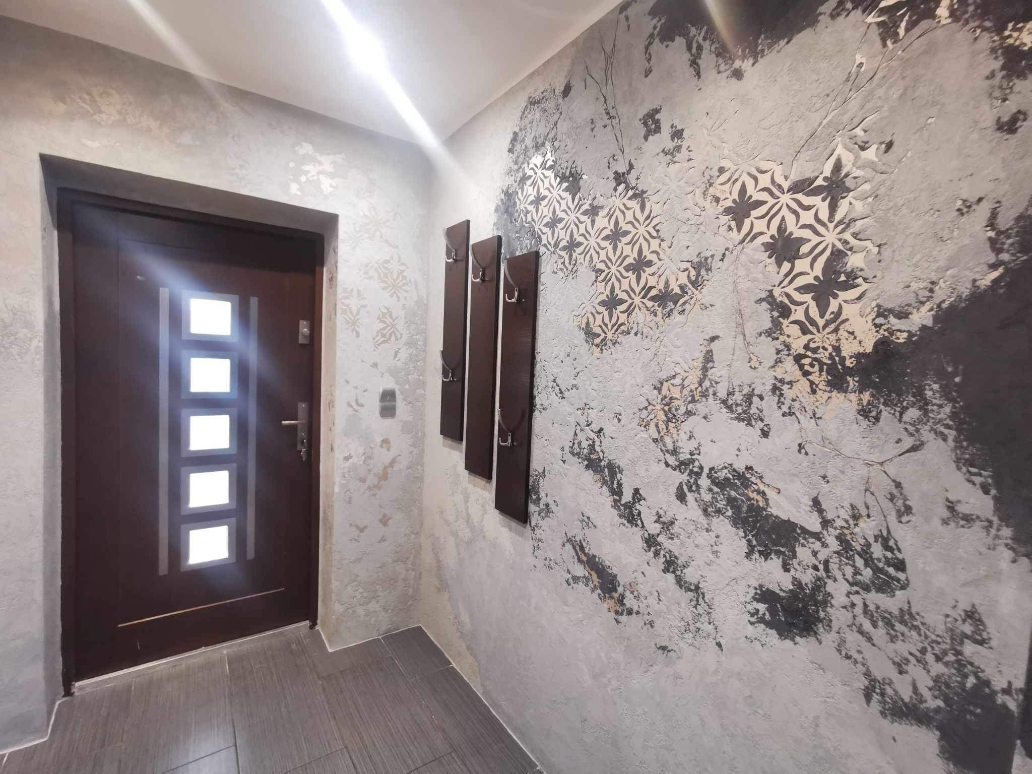 Hall z dekoracyjną ścianą w odcieniach szarości z motywem kwiatowym, brązowe wieszaki na ubrania i drzwi z przeszkleniami.