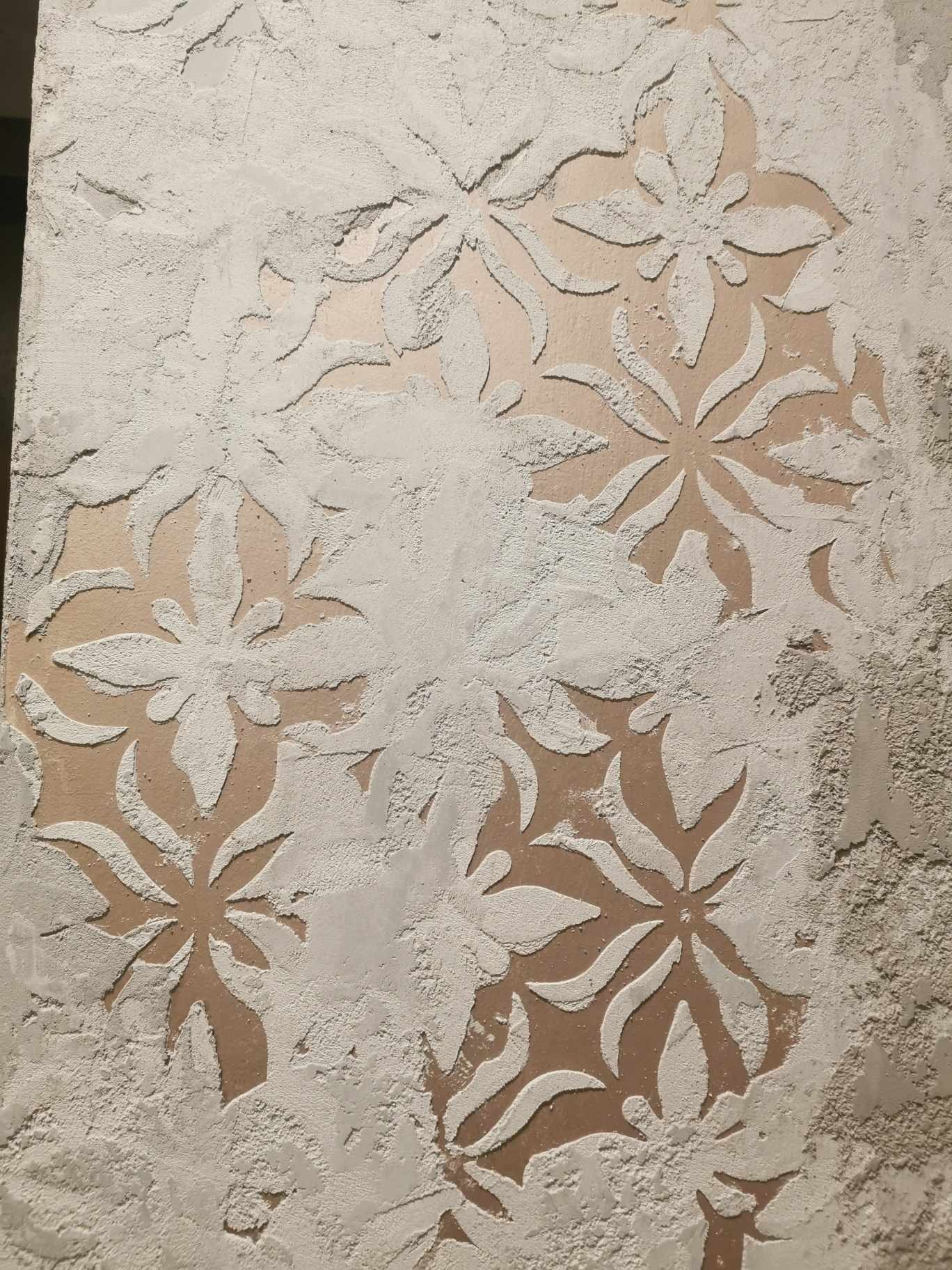 Ściana z dekoracyjną, kwiatową strukturą wykonaną z jasnej masy, z widocznymi niedoskonałościami i nierównościami na powierzchni.