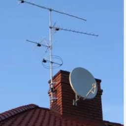 Antena telewizyjna i satelitarna zamontowane na kominie z czerwonej cegły, widoczne na tle błękitnego nieba i czerwonego dachu z dachówki.