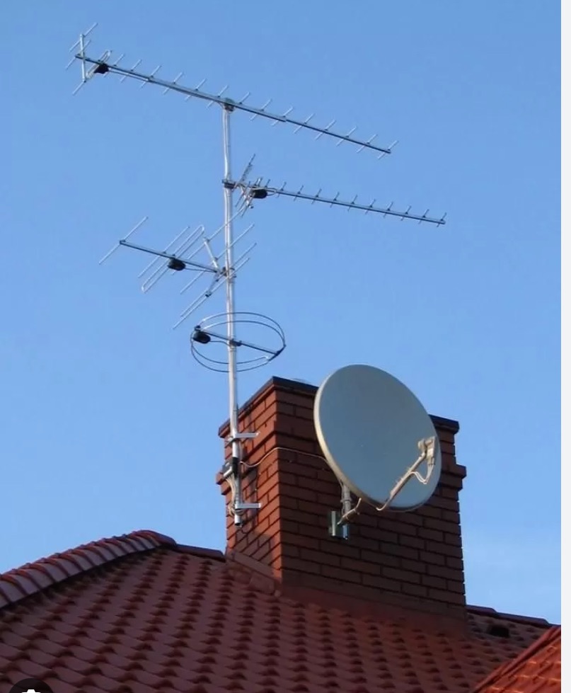 Antena telewizyjna i satelitarna zamontowane na kominie z czerwonej cegły, widoczne na tle błękitnego nieba i czerwonego dachu z dachówki.