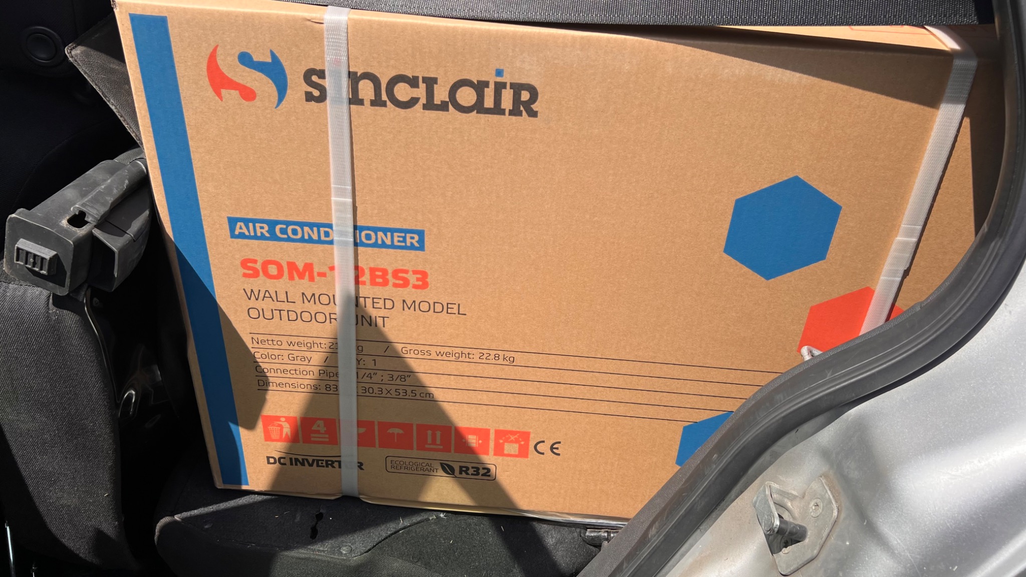 Karton klimatyzatora Sinclair SOM-12BS3, model do montażu na ścianie, jednostka zewnętrzna, przewożony w samochodzie. Widoczne dane techniczne i logo Sinclair.