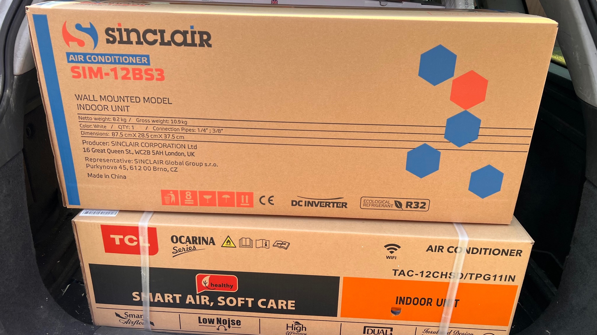 Dwa kartony klimatyzatorów Sinclair i TCL, umieszczone jeden na drugim w bagażniku samochodu. Widoczne specyfikacje techniczne i oznaczenia modeli.