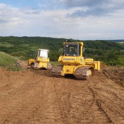 Bauma Rafał Dzido - Dwa żółte spychacze Komatsu na nierównym, brązowym terenie. W tle zielone wzgórza i błękitne niebo z obłokami. Jeden spychacz bliżej, drugi dalej, widoczny operator.