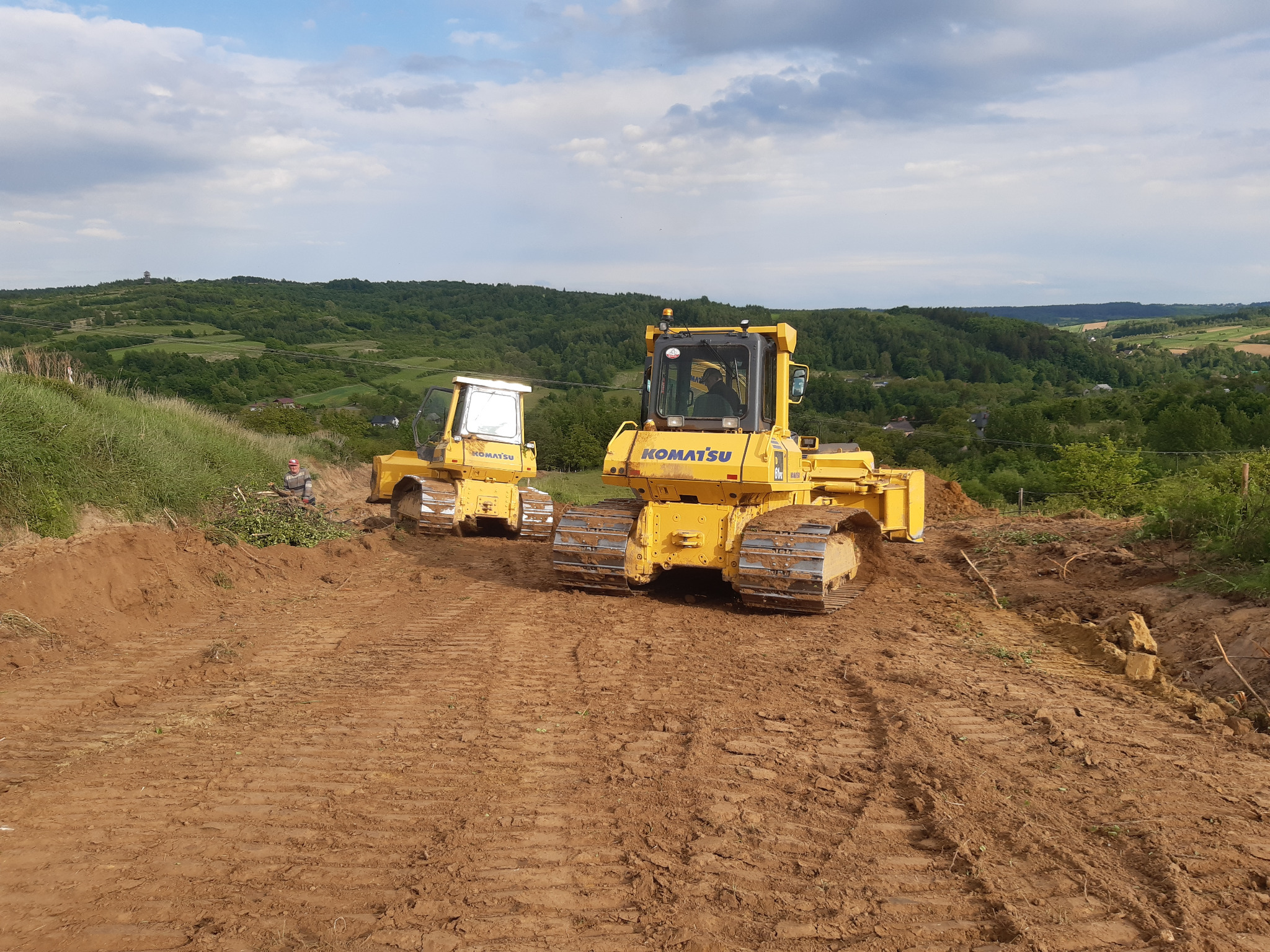 Dwa żółte spychacze Komatsu na nierównym, brązowym terenie. W tle zielone wzgórza i błękitne niebo z obłokami. Jeden spychacz bliżej, drugi dalej, widoczny operator.