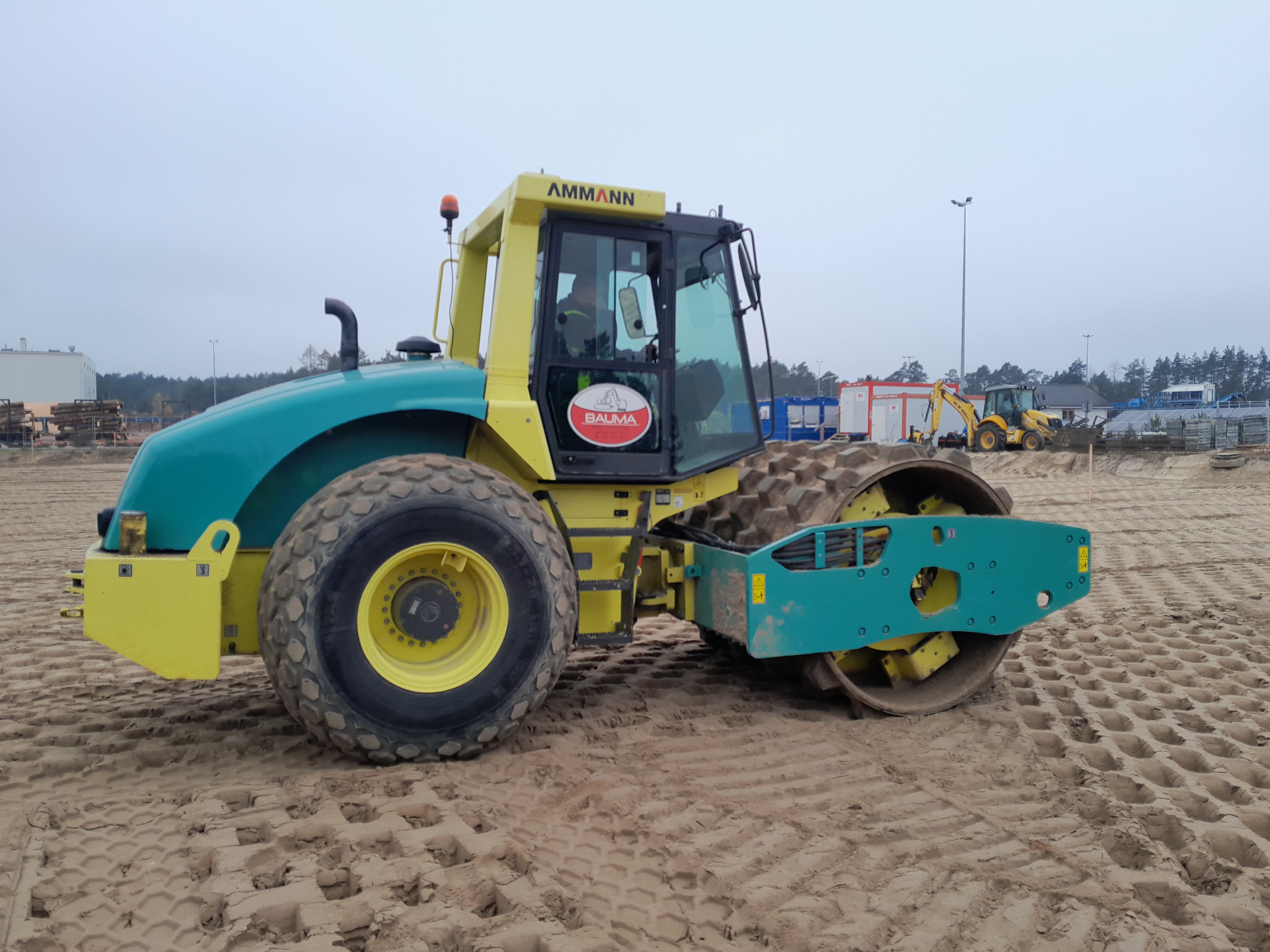 Żółto-turkusowy walec Ammann na piaszczystym placu budowy z widocznymi śladami po pracy, w tle budynki i koparka. Maszyna gotowa do utwardzania terenu pod inwestycję.