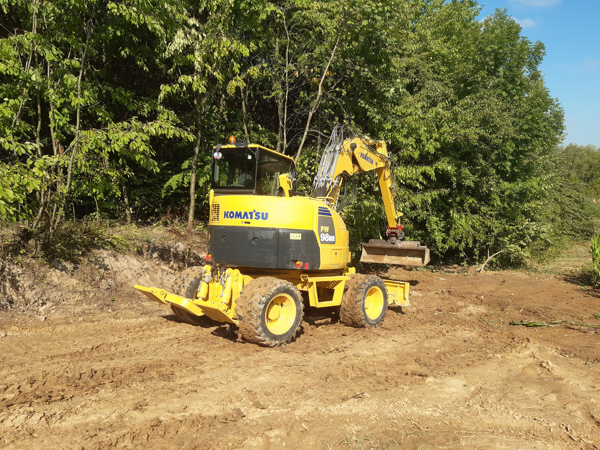 Żółta koparka kołowa Komatsu PW98MR na błotnistym terenie, z zielenią w tle i błękitnym niebem. Widoczne ślady opon w ziemi, sugerujące prace ziemne.