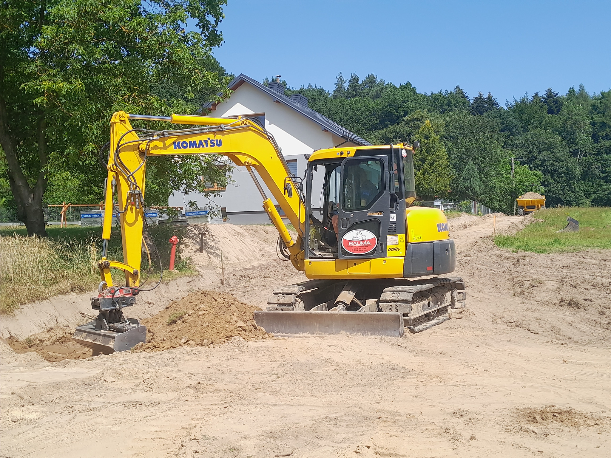 Żółta koparka Komatsu podczas pracy na piaszczystym terenie. W tle dom z ciemnym dachem i zieleń. Widoczny wykop i mała hałda ziemi.
