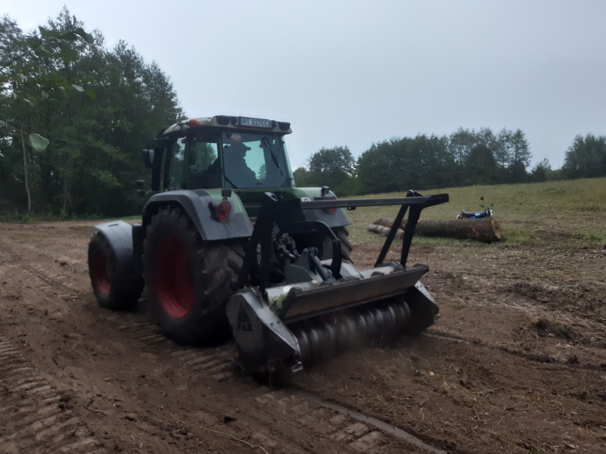 Ciągnik z glebogryzarką w trakcie niwelacji terenu. Widoczne ślady opon na ziemi i fragment zniwelowanego obszaru. W tle drzewa i pochmurne niebo.