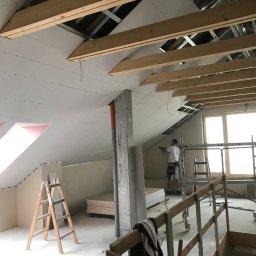 RENOVATION SQUAD - Montaż płyt kartonowo-gipsowych na skośnym poddaszu, widoczne drewniane belki stropowe i rusztowanie, pracownik w trakcie mocowania płyty, narzędzia pracy na podłodze.
