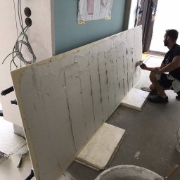 RENOVATION SQUAD - Mężczyzna nakłada szpachlę na płytę kartonowo-gipsową w trakcie prac wykończeniowych, widoczny kubeł z zaprawą, szpachelka i instalacja elektryczna w ścianie.