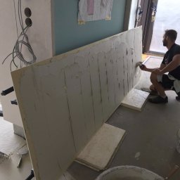 RENOVATION SQUAD - Mężczyzna w trakcie nakładania gładzi szpachlowej na dużą płytę kartonowo-gipsową w pomieszczeniu z odkrytą instalacją elektryczną i planami na ścianie.