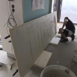 Przygotowanie płyty karton gips z nałożoną warstwą kleju, wspartej na podłodze i przytrzymywanej przez zaciski, widoczny pracownik przygotowujący się do montażu, wiadro z klejem i narzędzia...