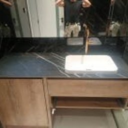 RENOVATION SQUAD - Łazienka: Umywalka nablatowa wpuszczona w czarny blat z marmurowym wzorem, złota bateria, szafka pod umywalką z drewnopodobnym fornirem, lustro w tle odbija osobę robiącą zdjęcie.