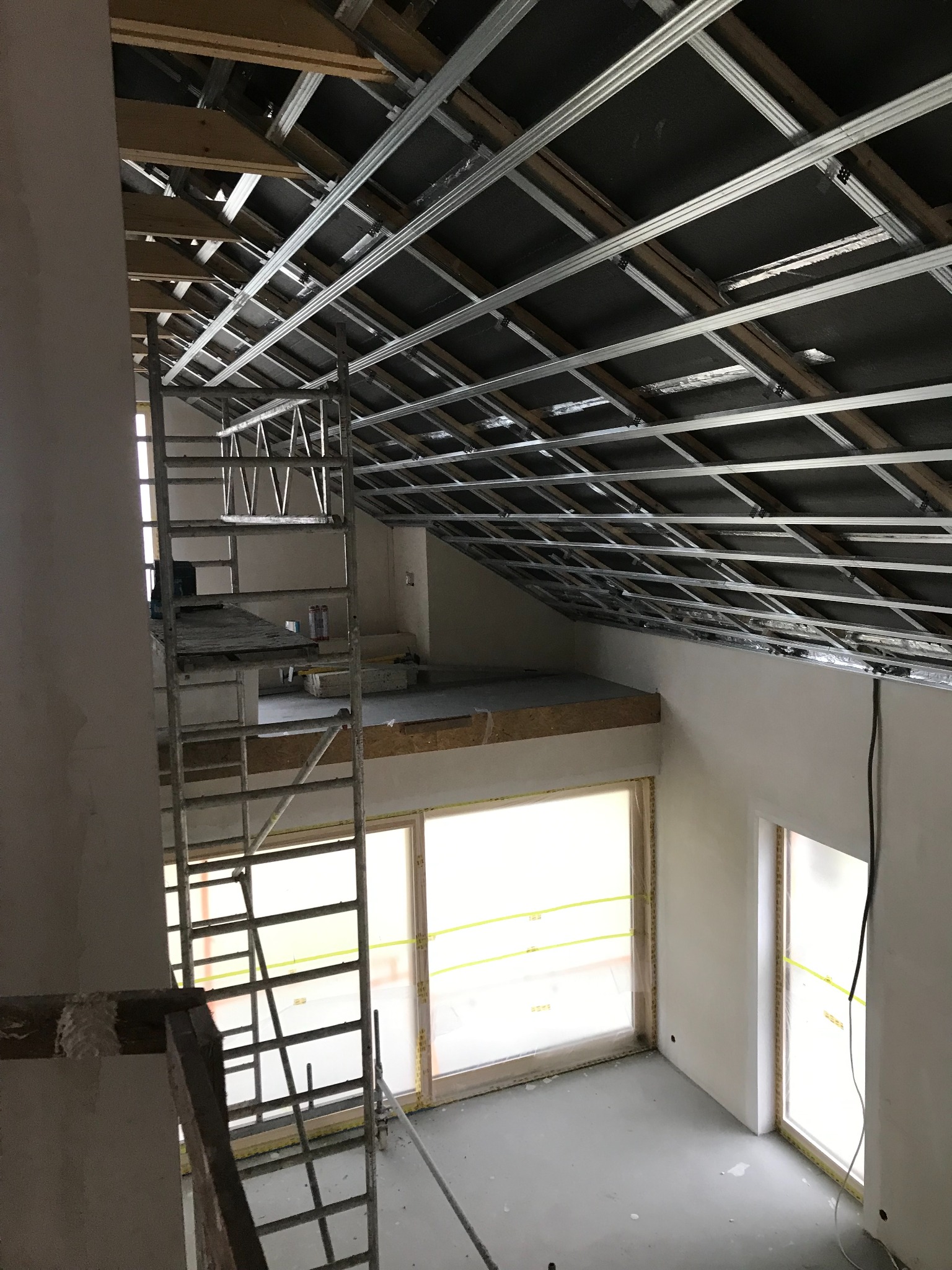 Szkielet sufitu podwieszanego z metalowych profili, widok z rusztowania w jasnym pomieszczeniu z oknami zabezpieczonymi folią.