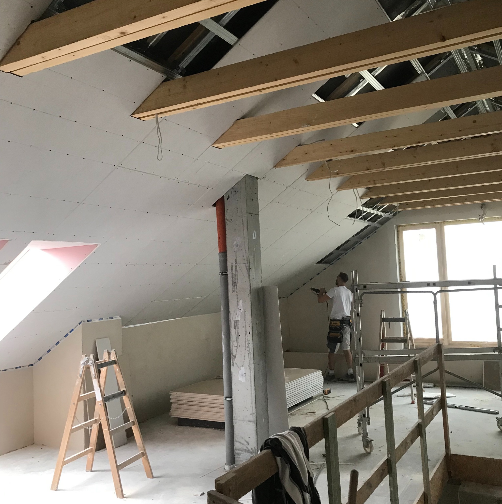 Montaż płyt kartonowo-gipsowych na skośnym poddaszu, widoczne drewniane belki stropowe i rusztowanie, pracownik w trakcie mocowania płyty, narzędzia pracy na podłodze.