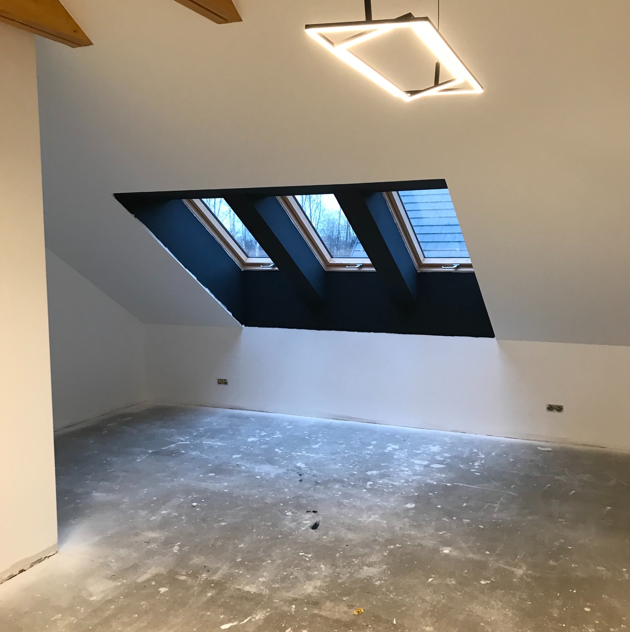 Poddasze z trzema oknami dachowymi w czarnej wnęce, surowa podłoga przygotowana do dalszych prac, nowoczesna lampa sufitowa.