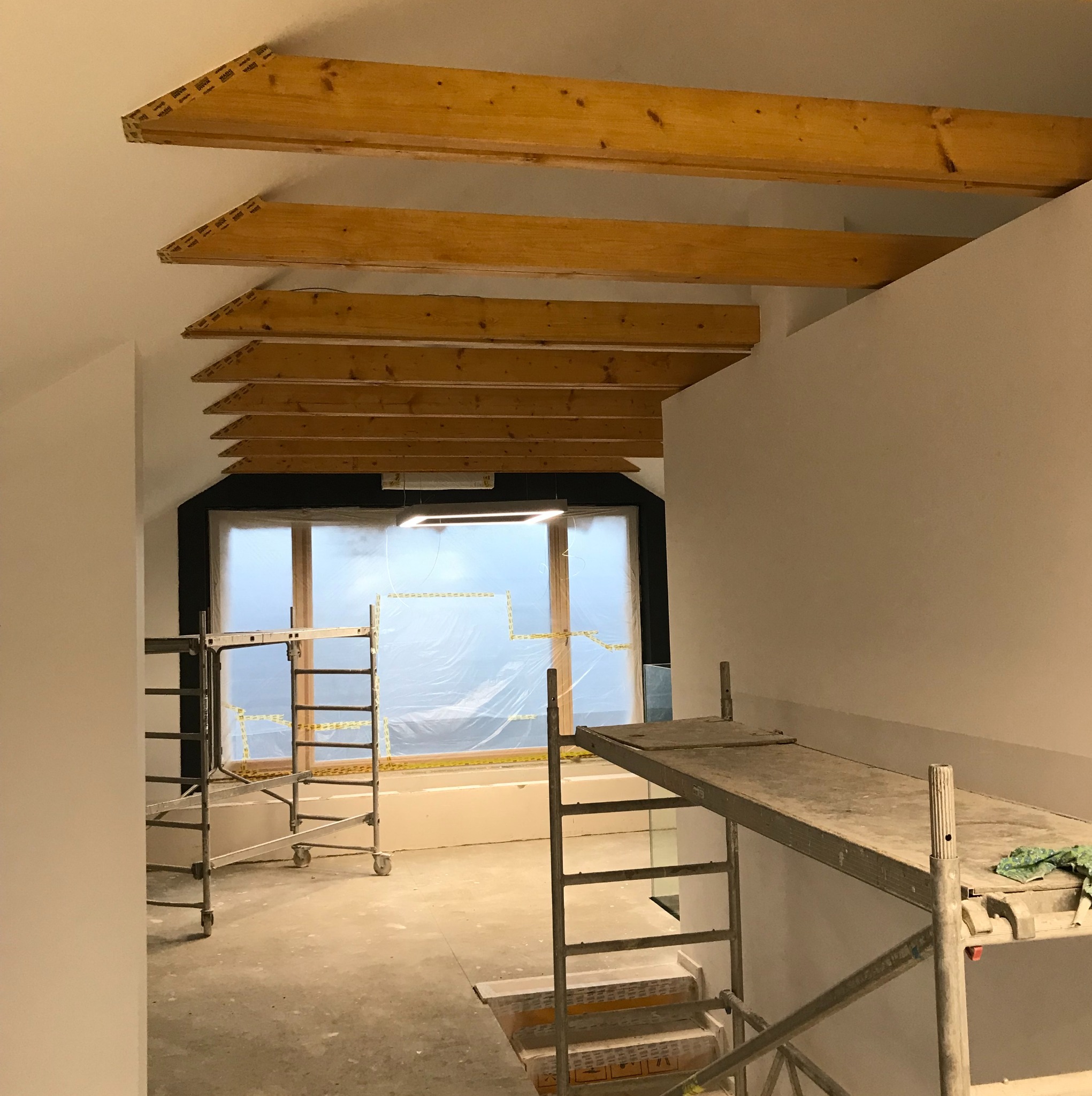 Wnętrze w trakcie prac wykończeniowych: widoczne drewniane belki stropowe, ściany przygotowane do malowania, okno zabezpieczone folią malarską oraz metalowe rusztowania.