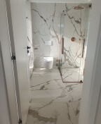 Nowoczesna łazienka z białymi płytkami imitującymi marmur, kabina prysznicowa typu walk-in ze złotą armaturą, podwieszana toaleta, widok zza otwartych drzwi.