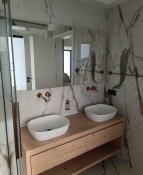 Nowoczesna łazienka z dwoma owalnymi umywalkami nablatowymi, drewnianą szafką z półkami, marmurowymi płytami na ścianach i lustrem. Baterie w kolorze różowego złota. Widoczny fragment kabiny...