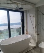 Biała, wolnostojąca wanna owalna na tle okna z widokiem na jezioro, ściany wyłożone marmurowymi płytami z wyraźnym, złotym żyłowaniem, ciemny sufit i minimalistyczna armatura.