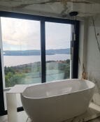 Nowoczesna, wolnostojąca wanna owalna przy dużym oknie z widokiem na jezioro i zielone wzgórza, marmurowe wykończenie ścian, czarna rama okienna i minimalistyczna lampa sufitowa.