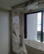 Wnętrze łazienki z wanną wolnostojącą, ściany wyłożone płytami imitującymi marmur z wyraźnym użyleniem, widok na zewnątrz przez duże okno.