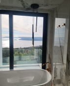 Nowoczesna łazienka z wolnostojącą wanną przy dużym oknie z widokiem na jezioro, minimalistyczna lampa wisząca, marmurowe wykończenie ścian, transparentna kabina prysznicowa.