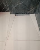 Białe płytki podłogowe z wąską, liniową kratką odpływową w kabinie prysznicowej typu walk-in, widoczne fragmenty ciemnej ściany z efektem marmuru.