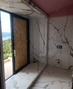 Wnętrze łazienki w trakcie prac wykończeniowych: białe płytki imitujące marmur na ścianach i posadzce, widoczne niedokończone połączenia na suficie, okno z czarną ramą zabezpieczone płytą OSB...