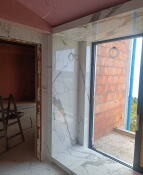 Wnętrze w trakcie prac wykończeniowych: ściany częściowo pokryte płytami imitującymi marmur, widoczna surowa ściana z cegły na zewnątrz okna balkonowego, różowa izolacja na suficie, brak podłogi...