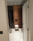 Nowoczesna łazienka z kabiną prysznicową typu walk-in, drewnianą ścianą z wbudowaną toaletą i białymi płytkami podłogowymi, widziana z korytarza przez otwarte białe drzwi.