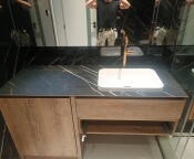 Łazienka: Umywalka nablatowa wpuszczona w czarny blat z marmurowym wzorem, złota bateria, szafka pod umywalką z drewnopodobnym fornirem, lustro w tle odbija osobę robiącą zdjęcie.